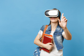 Young woman student in virtual reality goggles hold books touch something like push on button, pointing at floating virtual screen isolated on blue background. Education in school university college.