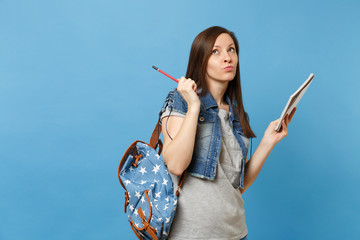 Portrait of young concerned woman student in denim clothes with backpack taking exam thinking about test hold notebook pencil isolated on blue background. Education in high school university college.