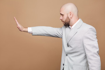 Profile side view portrait of middle aged bald bearded businessman in light gray suit standing with raised arms in stop gesture. indoor studio shot, isolated on light brown background.