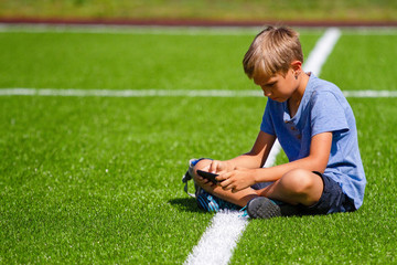 Boy sitting with cell phone sitting in empty football stadium field and looking video, typing,...
