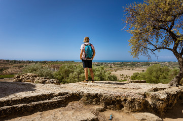 A tourist with a backpack watching a beautiful view during a sunny day (Valle dei Templi, Agrigento, Sicily, Italy) - obrazy, fototapety, plakaty