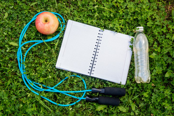 Bottle of water, notebook, apple and skipping rope on fresh green grass. Top view, copy space.
