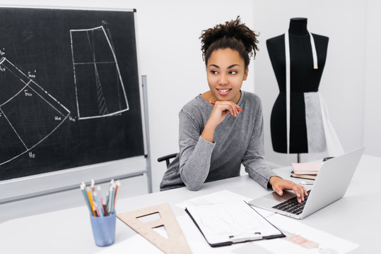 Portrait of young smiling African American designer planning project, typing on keyboard, sitting at workplace. University student learning fashion illustration using laptop computer for work. 