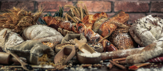 Bread background. Brown and white whole grain loaves wrapped in kraft paper composition on rustic dark wood with wheat ears scattered around. Baking and home bread making concept. Soft toning
