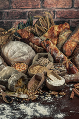 Bread background. Brown and white whole grain loaves wrapped in kraft paper composition on rustic dark wood with wheat ears scattered around. Baking and home bread making concept. Soft toning
