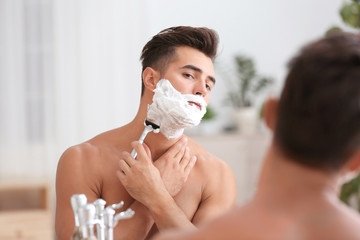 Young man shaving near mirror in bathroom
