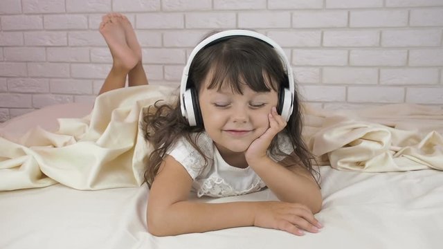 The child enjoys music. Charming little girl with headphones listening to music in bed.