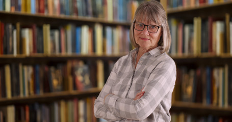 Confident senior woman with arms crossed standing indoors near bookshelves © Mark Adams