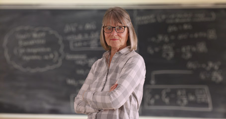 Confident senior woman teacher with arms crossed standing in front of chalkboard