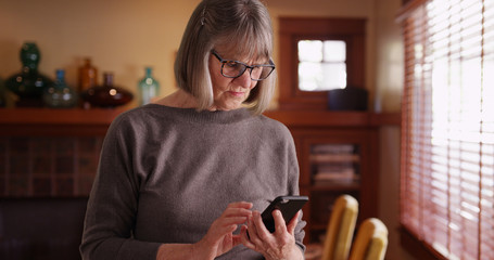 Serious senior woman using phone reading tragic news standing in living room