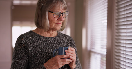 Somber old white woman contemplating and holding cup while looking out window
