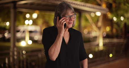 Lovely senior lady talking on smartphone with friend outdoors at night