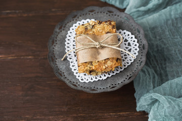 Homemade Chewy Granola Bars on a Tin Pedestal on a Wood Table; Granola Bar Wrapped in Parchment Paper and String; White Heart-Shaped Doily on Pedestal