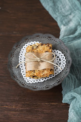 Fototapeta na wymiar Homemade Chewy Granola Bars on a Tin Pedestal on a Wood Table; Granola Bar Wrapped in Parchment Paper and String; White Heart-Shaped Doily on Pedestal