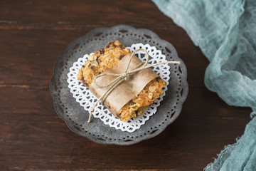 Homemade Chewy Granola Bars on a Tin Pedestal on a Wood Table; Granola Bar Wrapped in Parchment Paper and String; White Heart-Shaped Doily on Pedestal