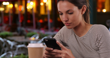 Young Caucasian lady with coffee cup using phone seated outdoors at restaurant
