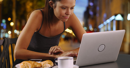 Young woman in Paris sitting outside using laptop and eating croissant