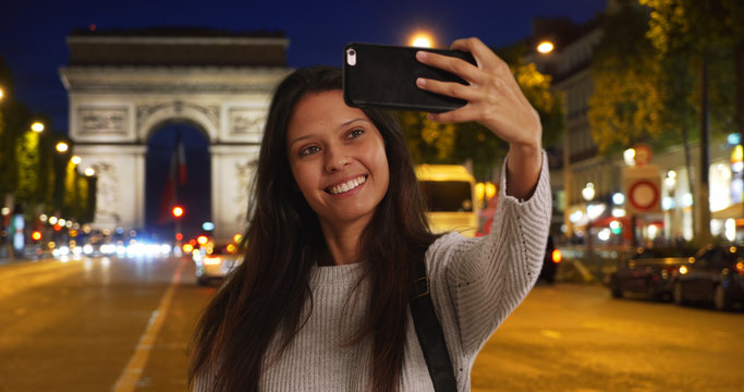 Beautiful female takes phone selfie at night with Arc de Triomphe behind her
