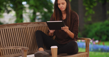 Millennial woman sitting on park bench reading with tablet computer