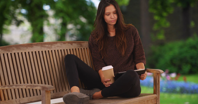 Millennial woman sitting on park bench reading on tablet and drinking coffee