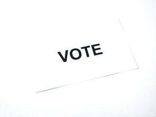 Photograph of a white piece of paper laying on a white background with the word vote on it in capital letters.
