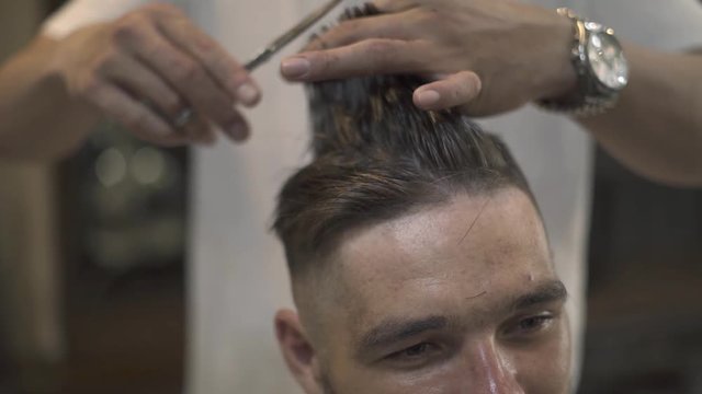 Face young man getting stylish hairdo in barbershop. Haircutter combing hair and cutting hair with hairdressing scissors in male salon. Close up hand hairdresser cutting wet male hair.