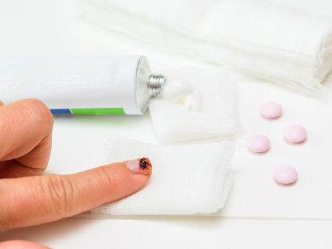 A knife-cut index finger of the left female hand, a tube of ointment and a bandage and pills on a white wooden table.