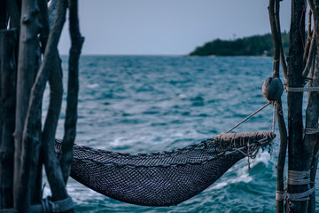 Brown net hammock hanging between the dry trees at the beach at Koh Raham. Photograph taken during dusk. The background is the sea waves splashing water and an island at horizon. 