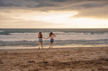 back portrait of two happy and attractive young women girlfriends holding hands on the beach running to the sea under beautiful sunset light enjoying summer holidays