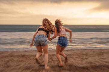 back portrait of two happy and attractive young women girlfriends holding hands on the beach running to the sea under beautiful sunset light enjoying summer holidays