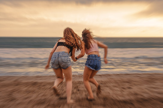 back portrait of two happy and attractive young women girlfriends holding hands on the beach running to the sea under beautiful sunset light enjoying summer holidays