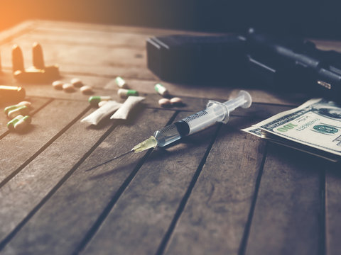 Selected focus on hypodermic syringe with blurred background of drugs, gun, bullet and bank note in dark room. Drug addicted concept.