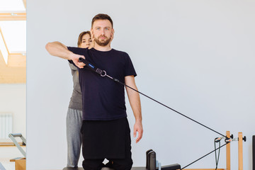 Brunette female instructor with cute two braids hairstyle consulting hansdome beard man workout pilates on reformer practice in pilates studio, working out indoor, correcting beginners, full length.