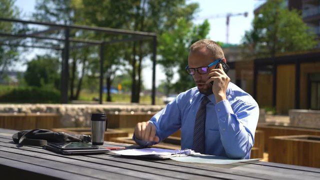 Angry businessman with documents talking on cellphone at city park
