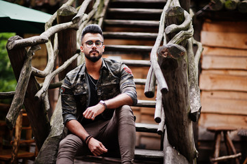 Awesome beautiful tall ararbian beard macho man in glasses and military jacket posed outdoor against wooden stairs.