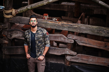 Awesome beautiful tall ararbian beard macho man in glasses and military jacket posed outdoor against wooden rustic house.