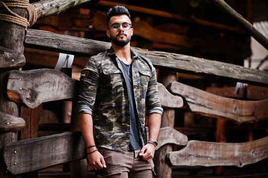 Awesome beautiful tall ararbian beard macho man in glasses and military jacket posed outdoor against wooden rustic house.