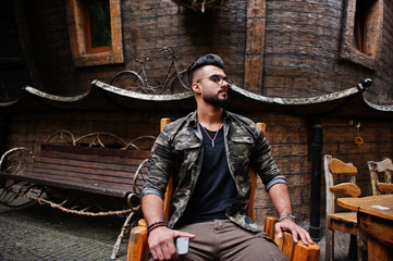 Awesome beautiful tall ararbian beard macho man in glasses and military jacket posed outdoor, sitting on chair against wooden house.