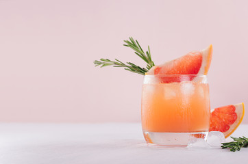 Refreshing cold citrus cocktail with ice cubes, green rosemary and slice grapefruit on soft light pink and white background.