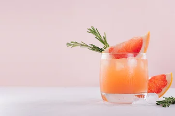 Gordijnen Verfrissende koude citruscocktail met ijsblokjes, groene rozemarijn en schijfje grapefruit op zachte lichtroze en witte achtergrond. © finepoints