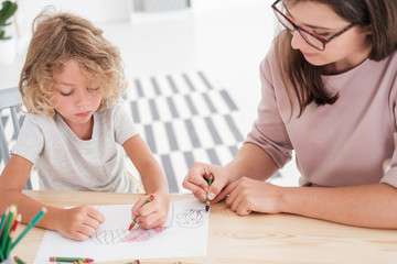 Little kid drawing a house using colorful crayons with his female, therapist during a meeting in...