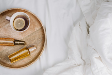 Flatlay of bed with white sheets, blankets and wooden tray with lemon tea and body or hair oils or serums, concept of morning or night skincare