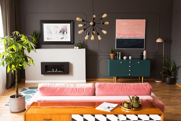 Plant next to pink couch in grey living room interior with poster above fireplace. Real photo