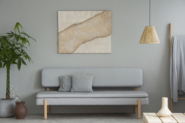 Poster above grey settee in simple living room interior with plant and lamp above table. Real photo
