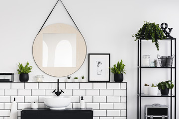 Round mirror and poster between plants in white and black bathroom interior with washbasin. Real...