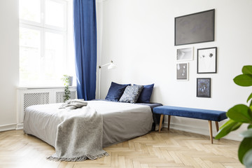 Bench next to bed with grey blanket in bright bedroom interior with posters and blue drapes. Real photo