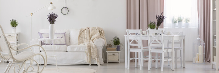 Panorama of white apartment interior with chairs at dining table next to sofa with blanket. Real photo