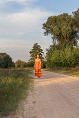 a girl in a rural summer long dress walks barefoot along a country sandy road at sunset in the...