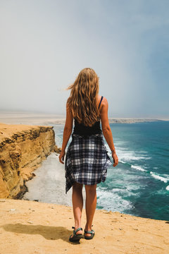 Summer. The girl (blonde) in shorts and top is standing with her back to the sea on a sandy cliff. Peru, Paracas