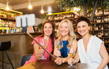 people, technology and lifestyle concept - women drinking wine and taking picture by smartphone on selfie stick at bar or restaurant